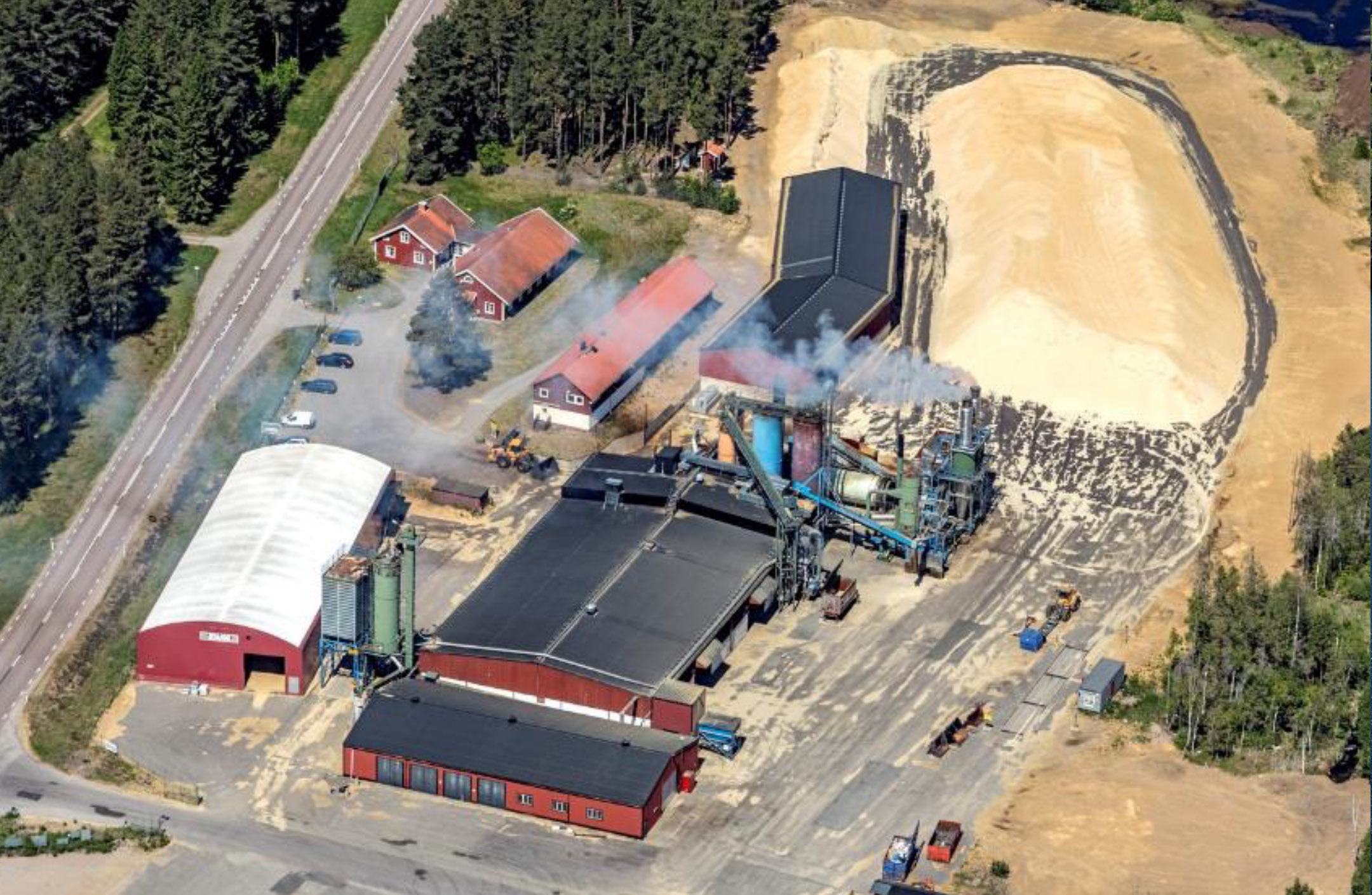 Flygbild pelletsfabriken i Främlingshem. Byggnader, spånhögar, riksvägen och vattenånga från torkprocessen